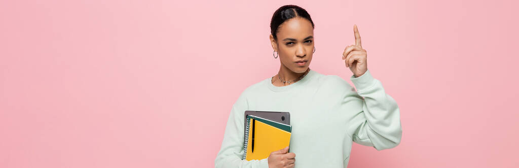 étudiant afro-américain intelligent tenant ordinateur portable et fournitures d'étude tout en ayant idée isolé sur rose, bannière  - Photo, image