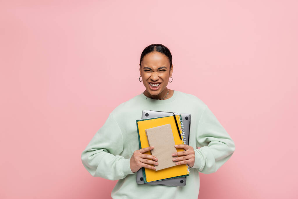 Αφροαμερικάνος φοιτητής με φούτερ κρατώντας φορητό υπολογιστή και να μελετήσει τις προμήθειες, ενώ χαμογελώντας απομονώνονται σε ροζ  - Φωτογραφία, εικόνα
