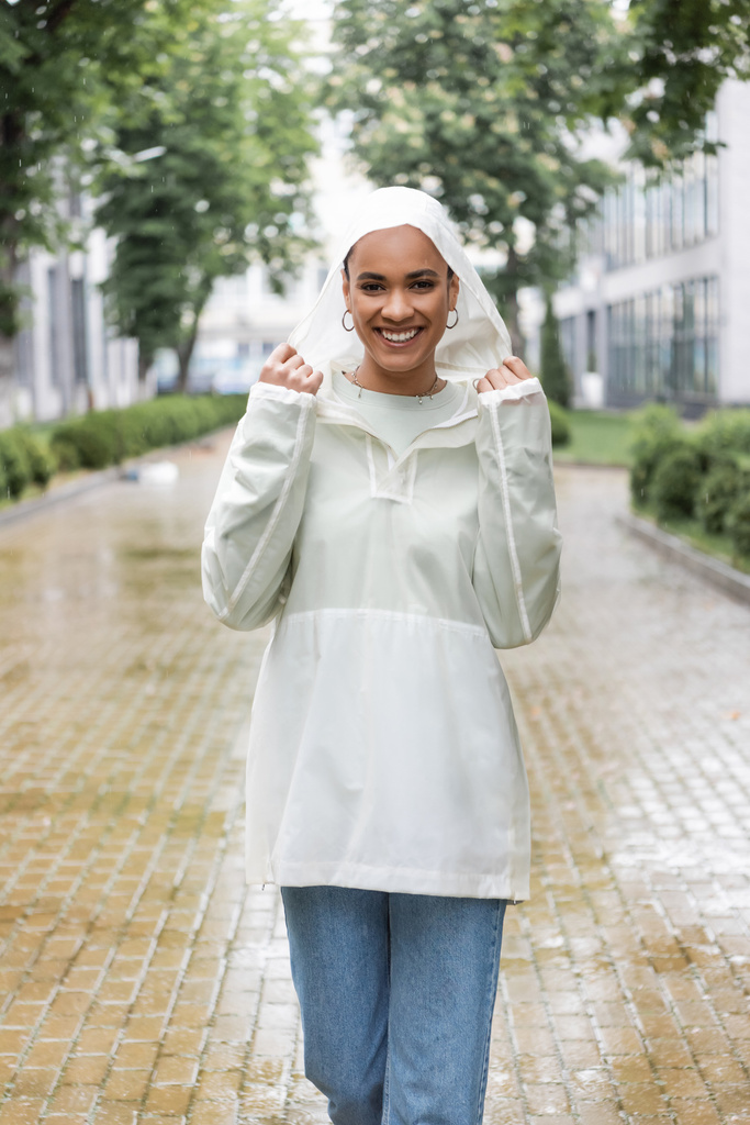屋外で雨の下に立って防水レインコートで陽気なアフリカ系アメリカ人女性  - 写真・画像
