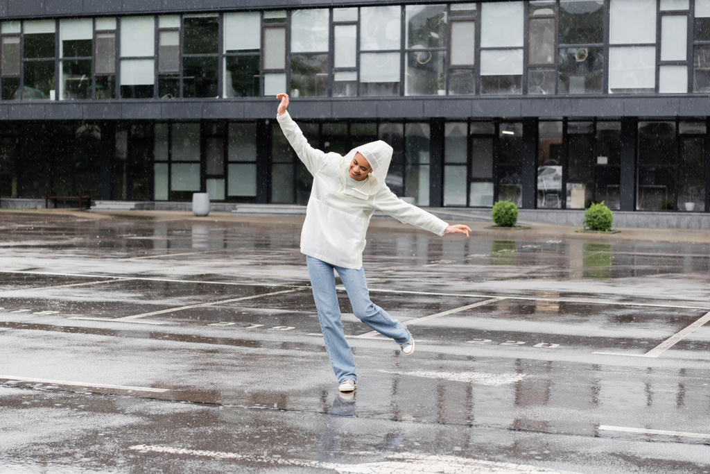 повна довжина щасливої афроамериканської жінки в водонепроникному плащі і джинсах, розважаючись під час дощу
  - Фото, зображення