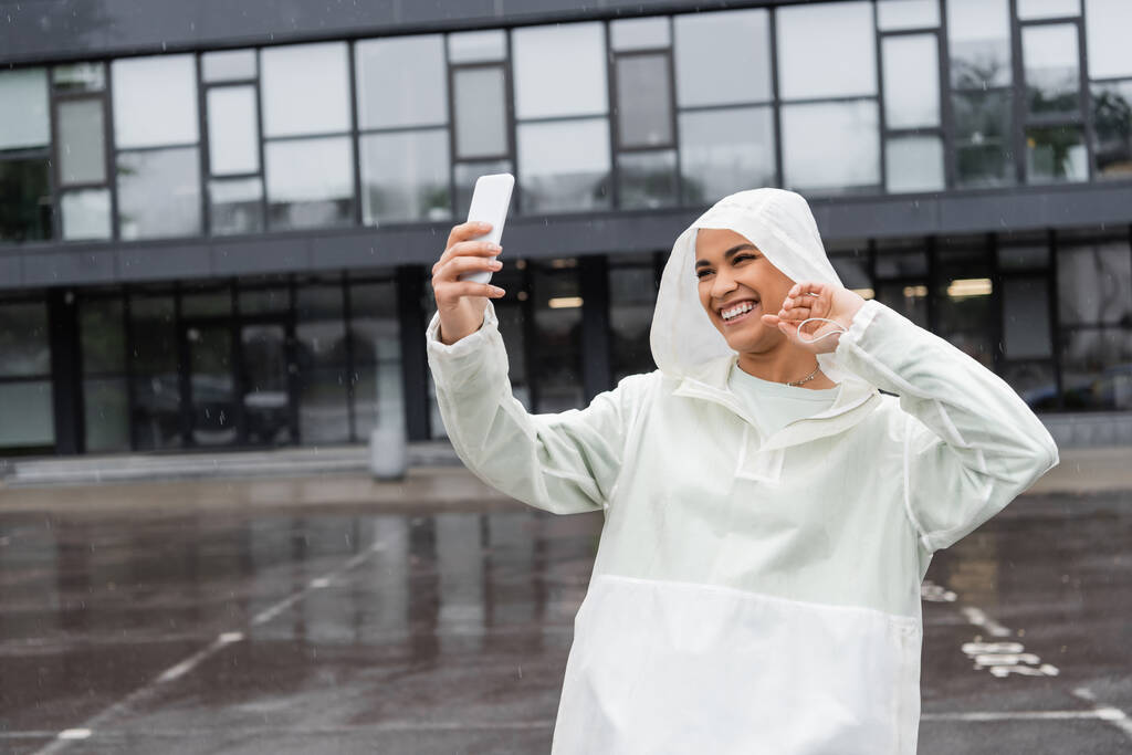 насолоджена афроамериканська жінка в водонепроникному плащі бере селфі на смартфон під час дощу
  - Фото, зображення
