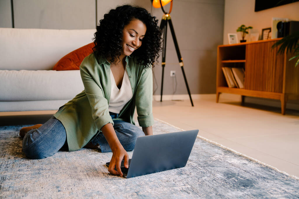 Fröhliche junge Afrikanerin benutzt Laptop, während sie zu Hause auf dem Boden sitzt - Foto, Bild