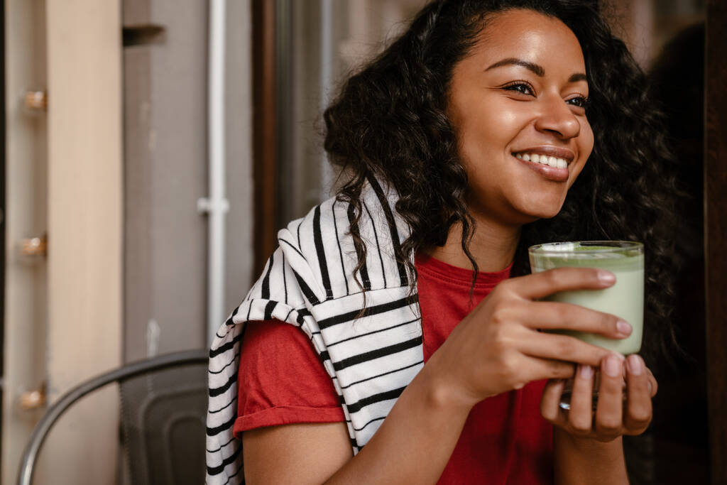 Nuori musta nainen hymyilee ja juo matcha latte istuessaan kahvilassa ulkona - Valokuva, kuva