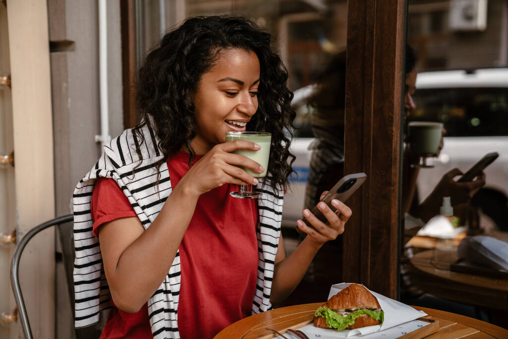 Молодая черная женщина пользуется мобильным телефоном и обедает в кафе на открытом воздухе - Фото, изображение