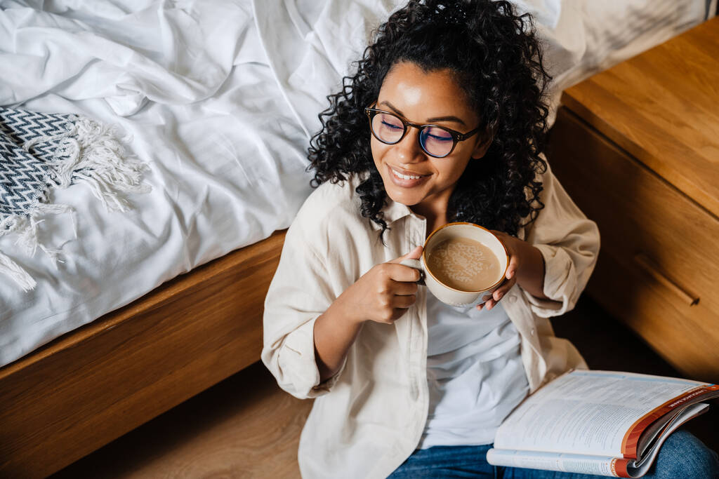 アフリカ系アメリカ人の若い女性が寝室で運動本を勉強しながらコーヒーを飲む - 写真・画像