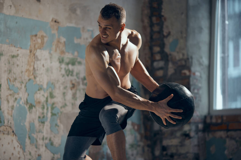 Trening całego ciała. Młody muskularny mężczyzna z mocną, ulgą sylwetki trening bez koszulki z piłką fitness w pomieszczeniach. Ciało potu. Pojęcie sportowego stylu życia, pielęgnacji ciała, fitness, hobby, zdrowia - Zdjęcie, obraz