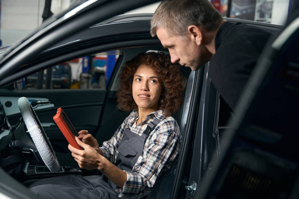 Multiraciale vrouwelijke auto reparateur zit in de auto met een apparaat en communiceren met de klant, een man van middelbare leeftijd in casual kleding - Foto, afbeelding