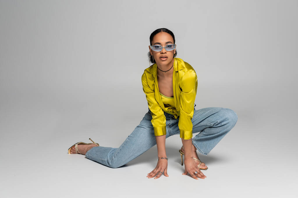 pełna długość stylowy afrykański amerykański model w niebieskich okularach przeciwsłonecznych pozowanie podczas stojąc na kolanie na szarym tle  - Zdjęcie, obraz