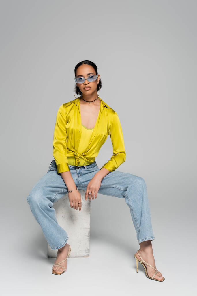 pełna długość afrykańskiego modelu amerykańskiego w stylowych okularach przeciwsłonecznych i stroju siedzącego na betonowej kostce podczas pozowania na szarości  - Zdjęcie, obraz