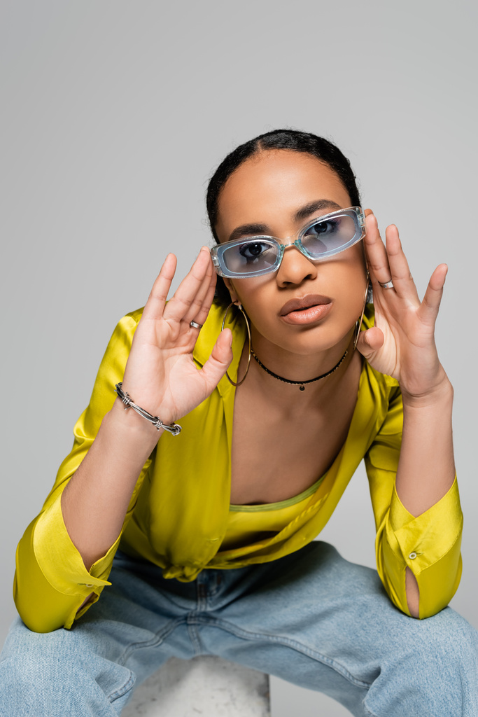 портрет молодої афроамериканської моделі, що регулює стильні сонцезахисні окуляри, дивлячись на камеру ізольовано на сірому
  - Фото, зображення
