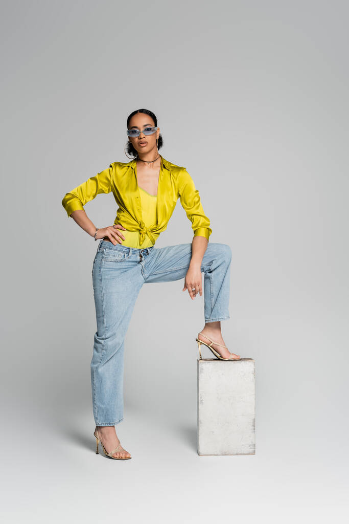 comprimento total do modelo americano africano confiante pisando no cubo de concreto enquanto posando com a mão no quadril no cinza  - Foto, Imagem