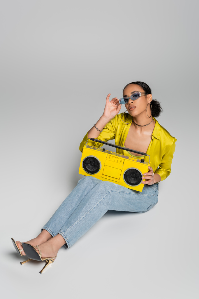 vue grand angle de femme afro-américaine élégante ajustant les lunettes de soleil bleues et assise avec boombox rétro sur gris  - Photo, image