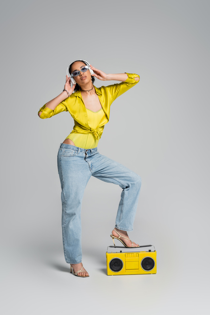 ワイヤレスヘッドフォンでアフリカ系アメリカ人女性の完全な長さ灰色の黄色のboomboxの近くで音楽を楽しむ  - 写真・画像
