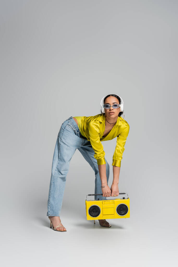πλήρες μήκος της κομψής αφρικανικής Αμερικής γυναίκα σε ασύρματα ακουστικά κρατώντας κίτρινο κουτί boombox σε γκρι  - Φωτογραφία, εικόνα
