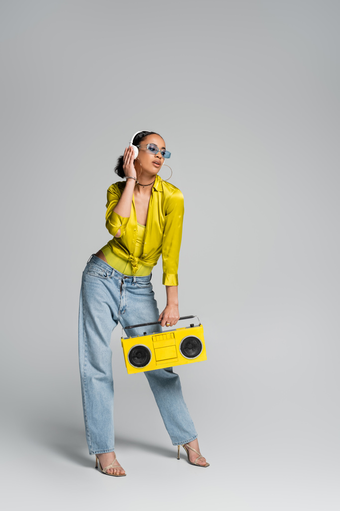 ワイヤレスヘッドフォンでブルネットアフリカ系アメリカ人モデルの完全な長さ灰色で黄色のboomboxを保持  - 写真・画像