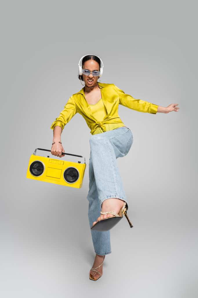 pełna długość emocjonalnego afrykańskiego modelu w słuchawkach bezprzewodowych trzymających żółty boombox i pozujących na szary  - Zdjęcie, obraz