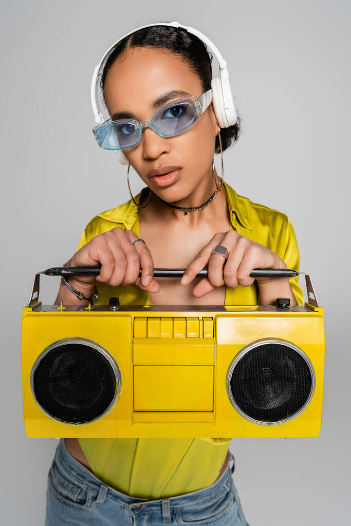 jolie femme afro-américaine écoutant de la musique dans des écouteurs sans fil et tenant boombox jaune isolé sur gris  - Photo, image