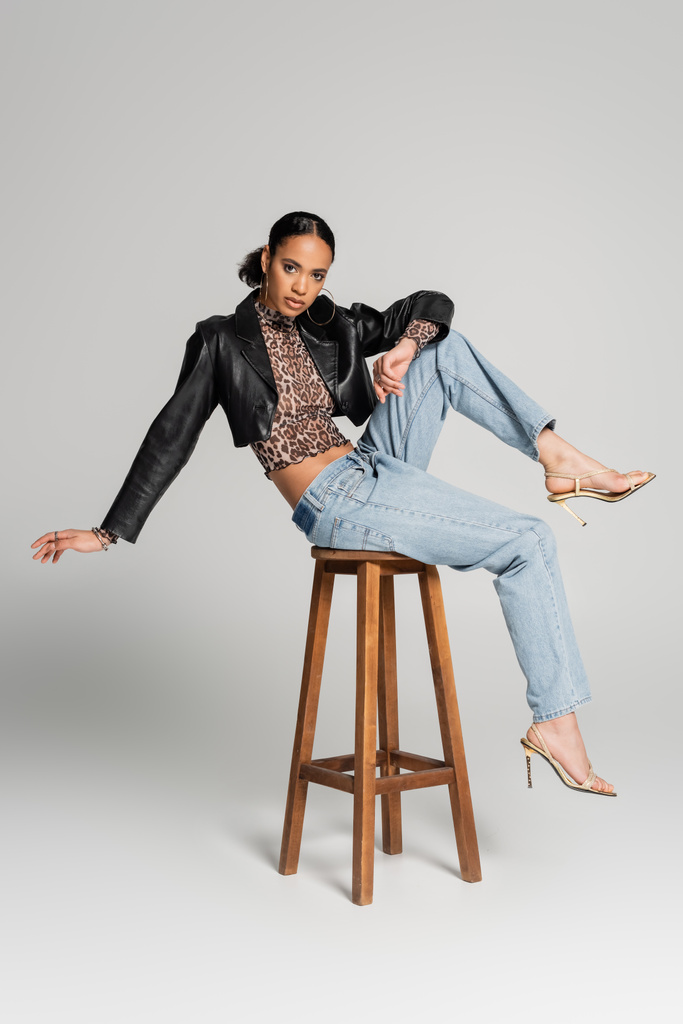полная длина молодой африканской американской модели в обрезанной куртке и сандалиях на высоких каблуках, позирующих на высоком стуле на сером - Фото, изображение