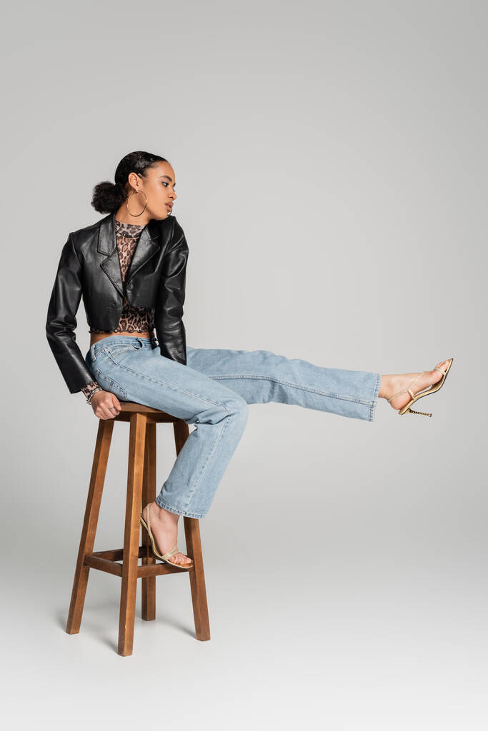 full length of young african american model in cropped jacket and jeans sitting on high stool on grey - Photo, Image