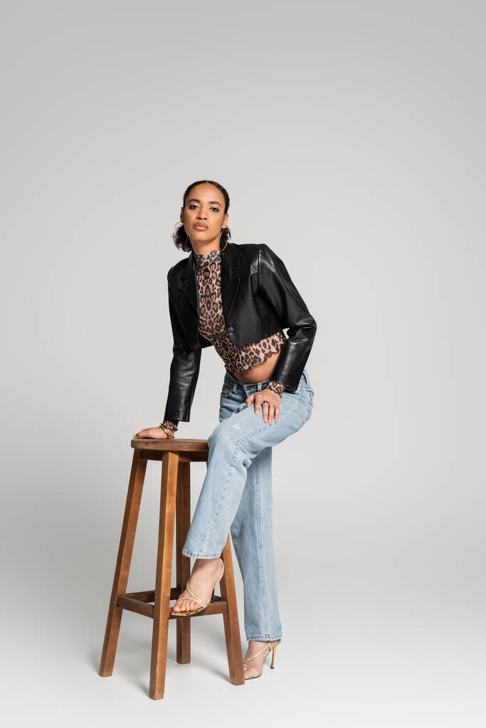 longitud completa de elegante modelo afroamericano en chaqueta recortada y pantalones vaqueros de pie cerca de la silla alta de madera en gris - Foto, Imagen