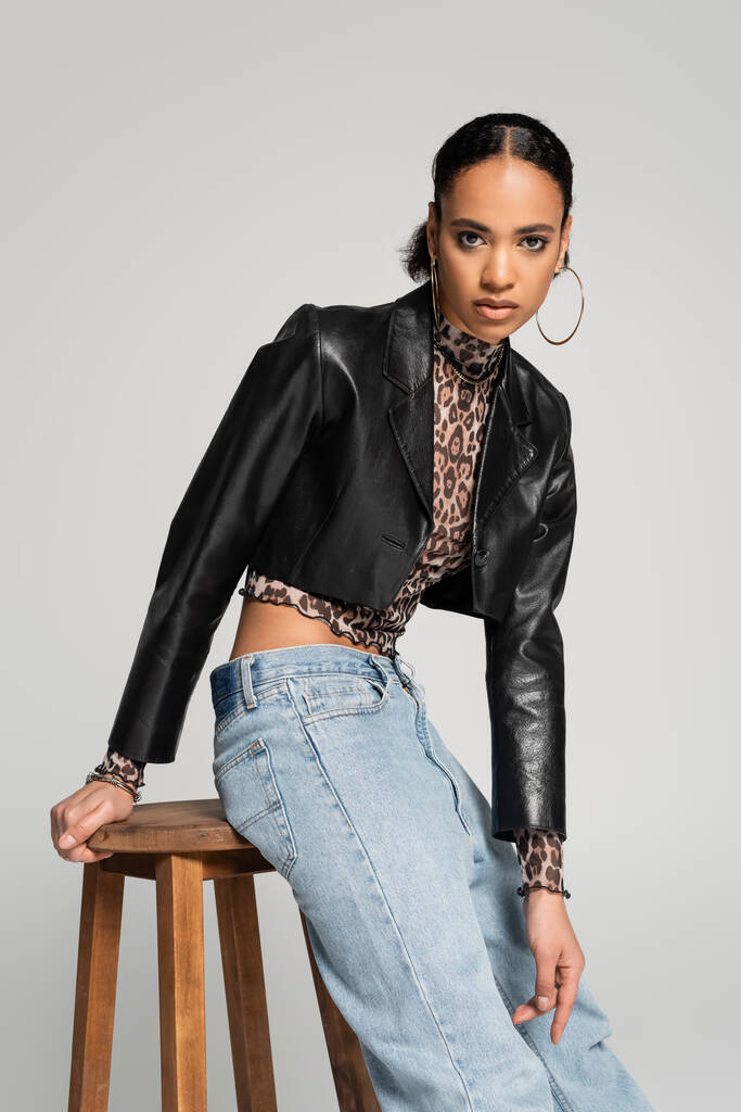 jovem modelo afro-americano na elegante jaqueta recortada e jeans posando perto de cadeira alta de madeira isolada em cinza - Foto, Imagem