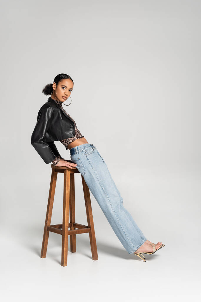 на всю длину модной африканской американки в обрезанной куртке и джинсах опирающейся на деревянный высокий стул на сером - Фото, изображение