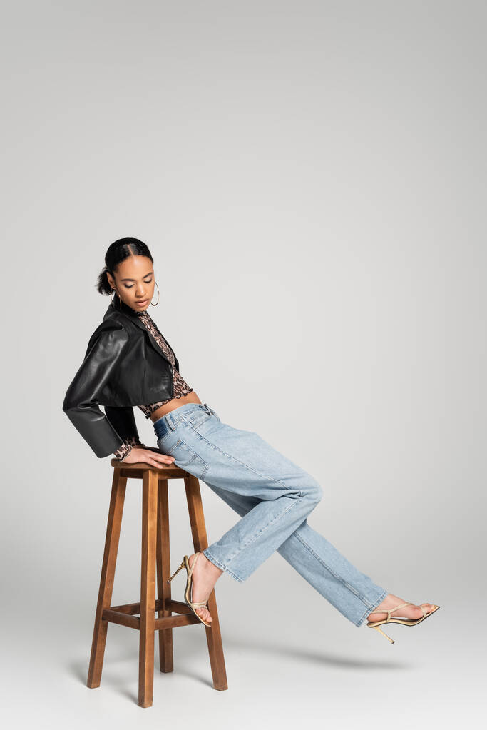 full length of stylish african american model in cropped jacket and jeans leaning on wooden high chair on grey - Photo, Image