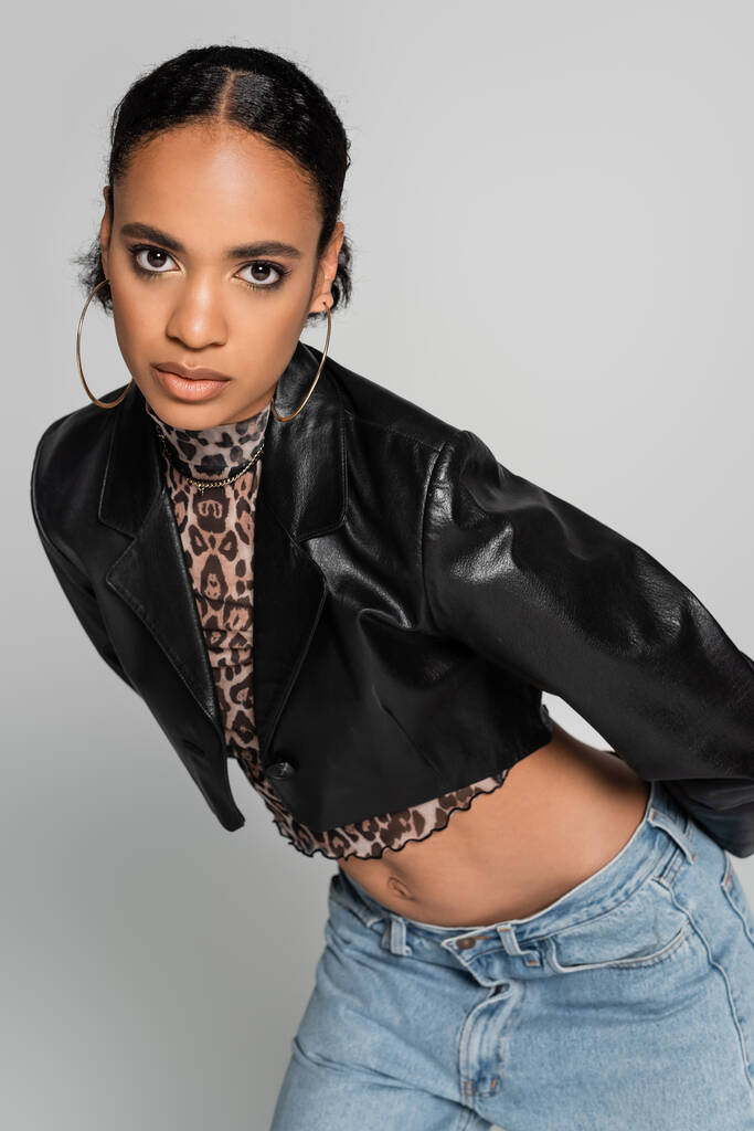 модна афро-американська модель в обрізаному піджаку і сережках кільця позує ізольовано на сірому
 - Фото, зображення