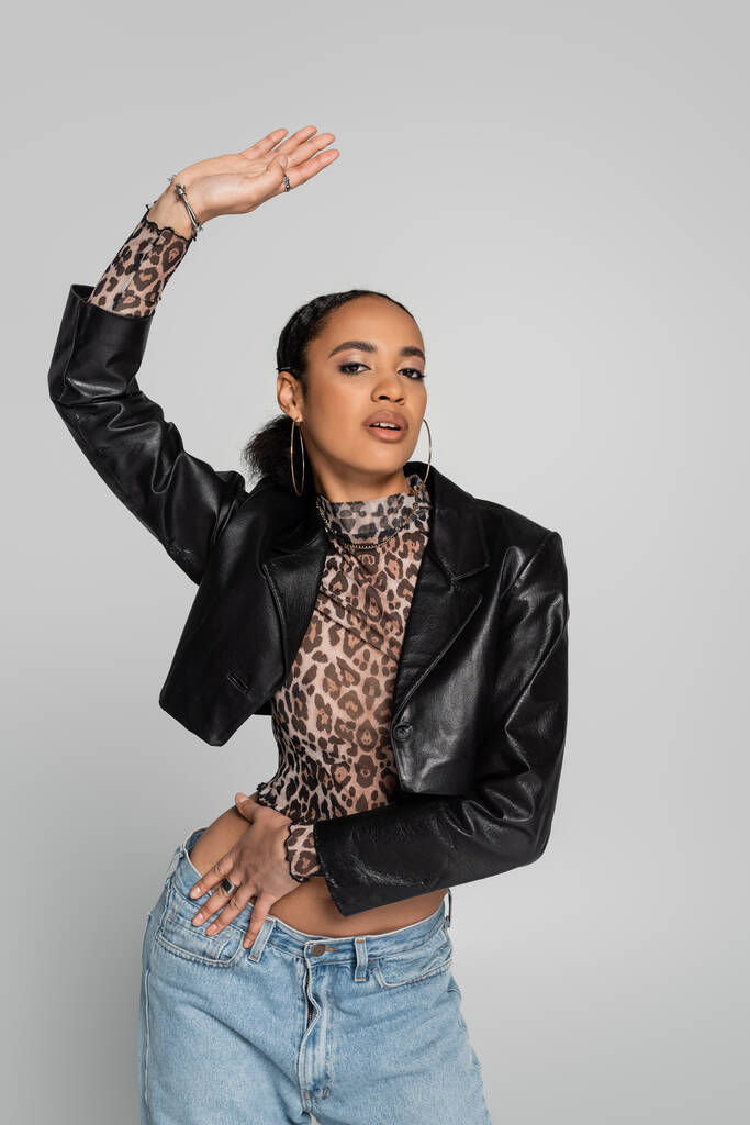 fashionable african american woman in black cropped jacket and top with animal print posing with raised hand isolated on grey  - Photo, Image