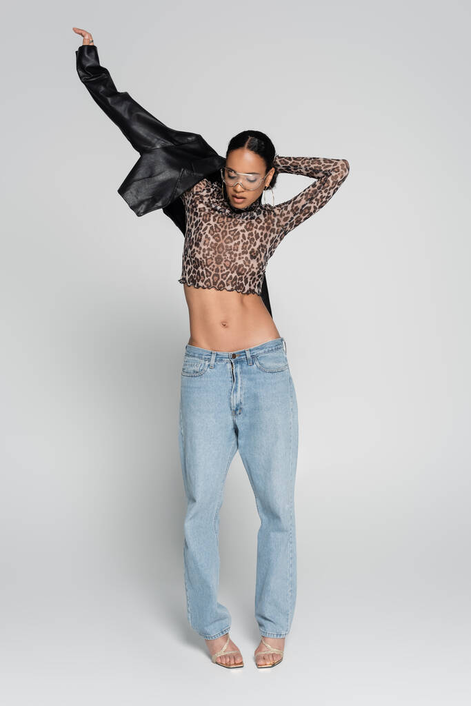 full length of stylish african american woman in crop top with animal print and sunglasses wearing leather jacket on grey  - Photo, Image