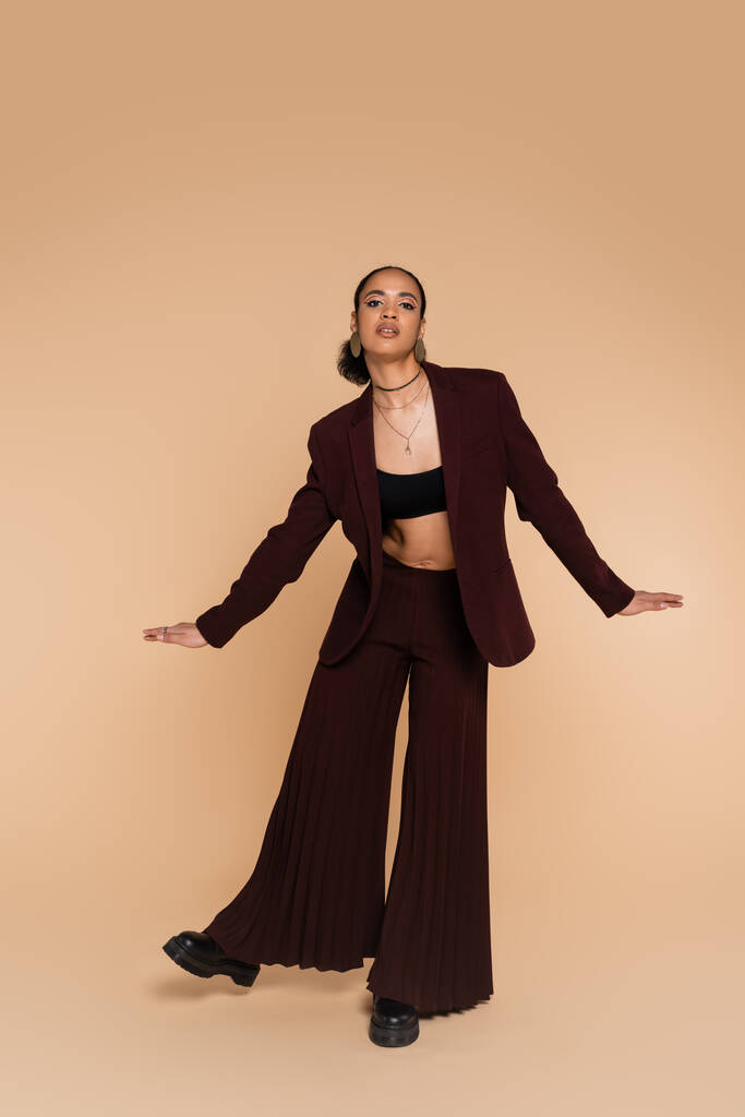 full length of young african american woman in stylish suit with wide pants gesturing while posing on beige  - Photo, Image