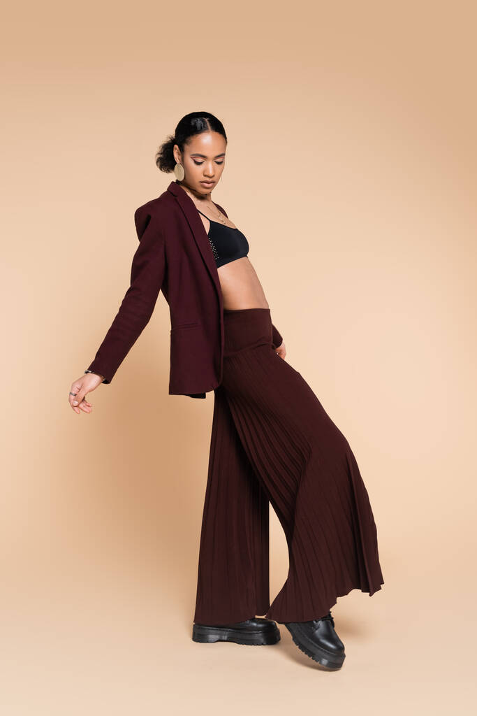longitud completa de la joven afroamericana en la parte superior de la cosecha y elegante traje marrón con pantalones de pierna ancha posando en beige  - Foto, imagen