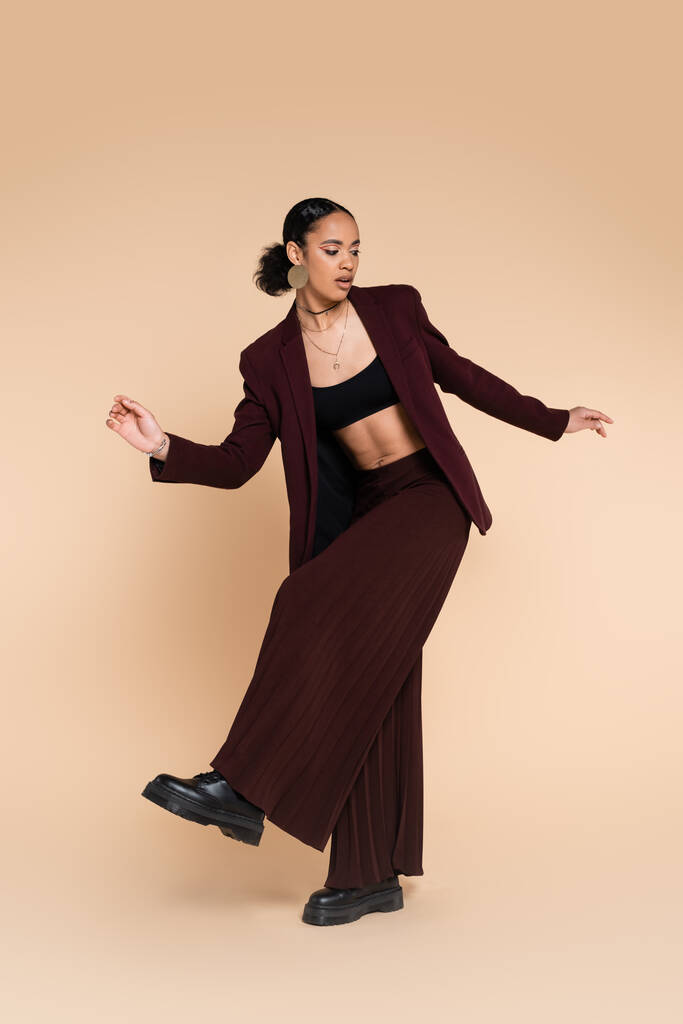 full length of african american woman in trendy maroon suit with wide leg pants posing on beige  - Photo, Image