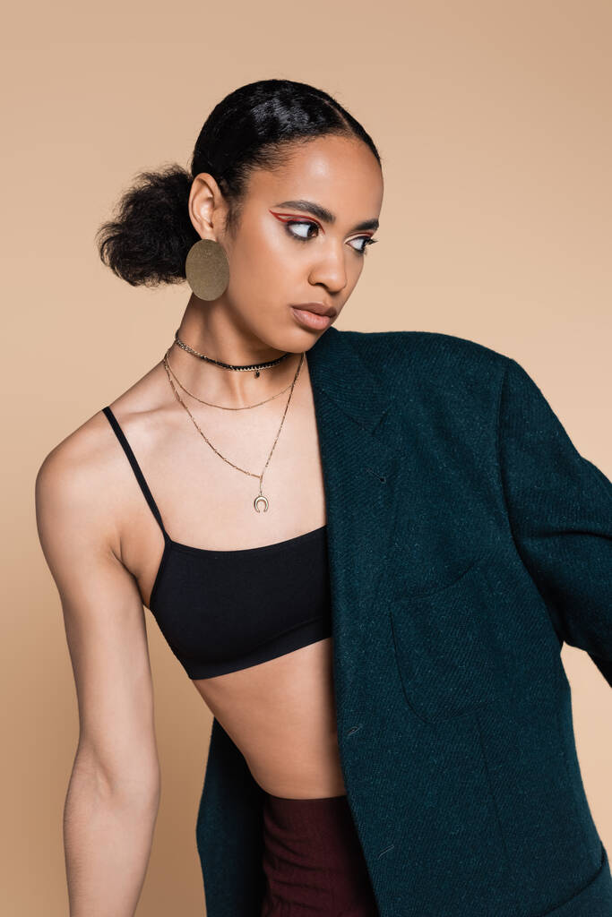 young african american model in crop top with dark blue oversized blazer on shoulder posing isolated on beige  - Photo, Image