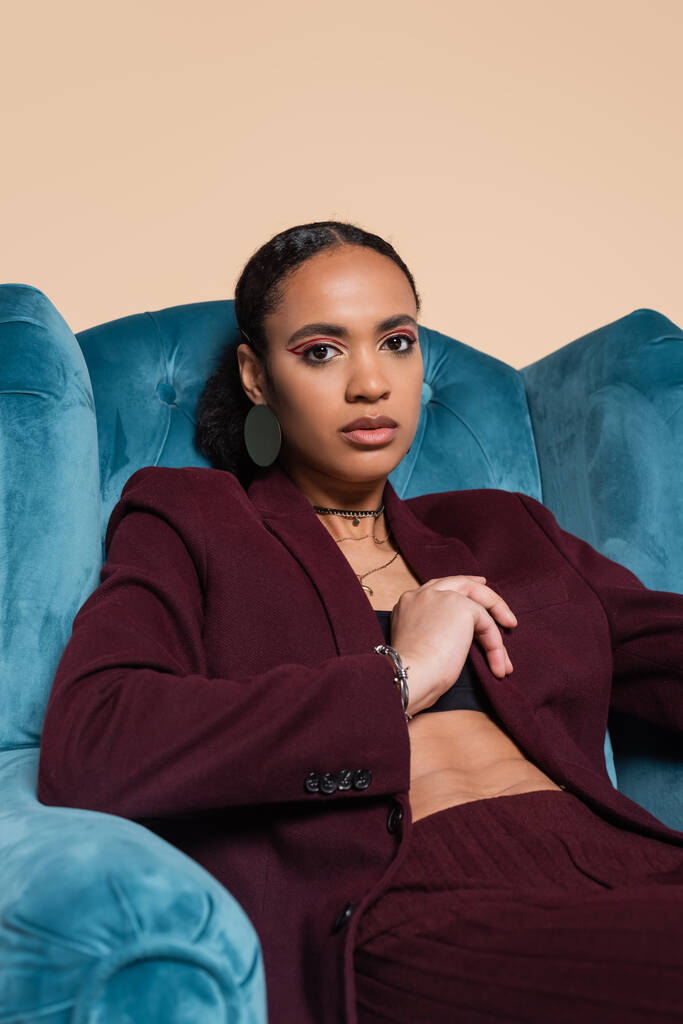 pretty african american model in maroon suit sitting on blue velvet armchair isolated on beige  - Photo, Image