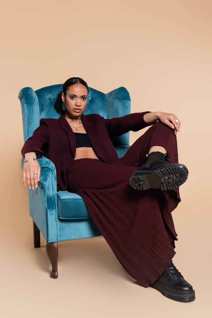 Afro-Amerikan modeli kestane rengi ceketli ve geniş pantolonlu bej renkli mavi kadife koltukta oturuyor.  - Fotoğraf, Görsel