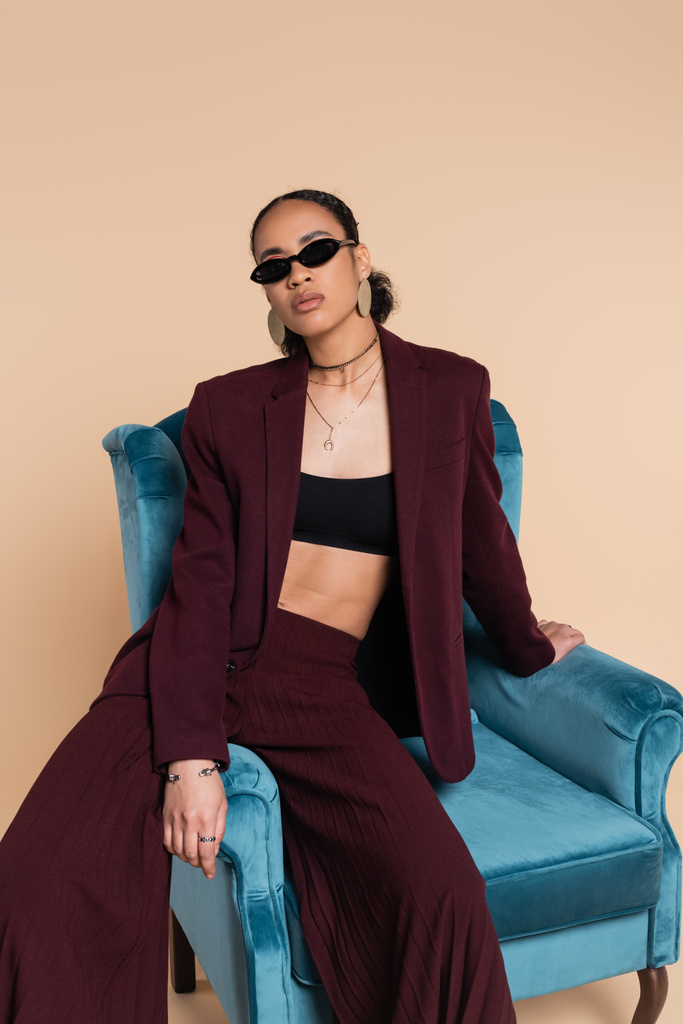 african american model in burgundy suit and stylish sunglasses sitting on blue velvet armchair on beige  - Photo, Image