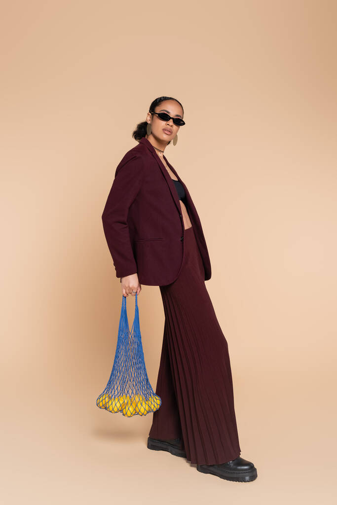 pełna długość stylowa Afroamerykanka w burgundowym garniturze i okularach przeciwsłonecznych trzymając torebkę z cytrynami na beżu   - Zdjęcie, obraz