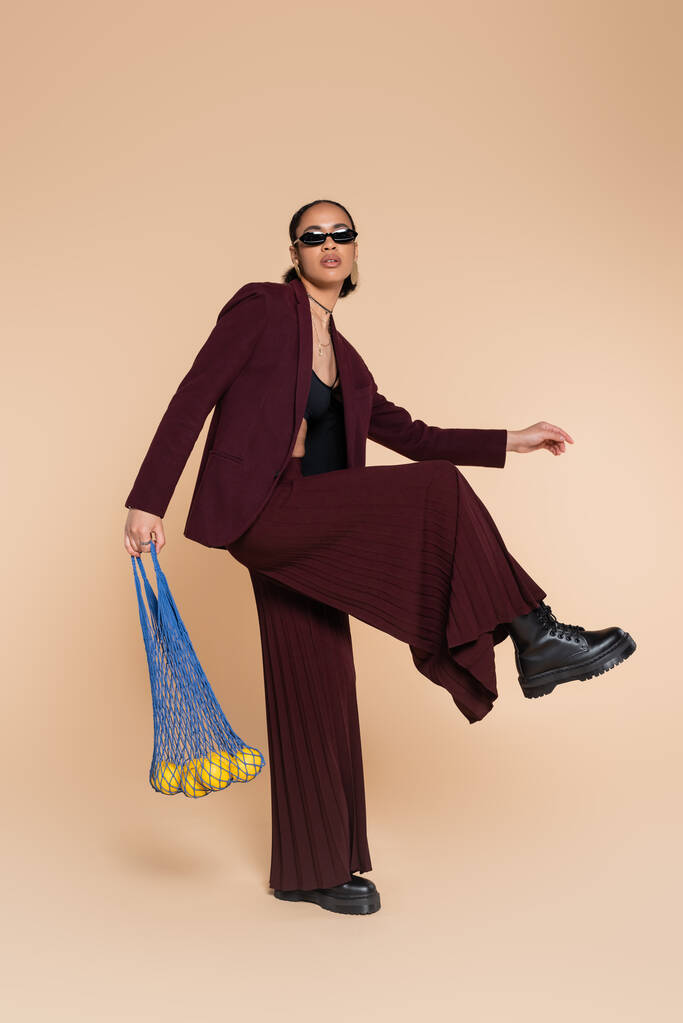 volledige lengte van stijlvolle Afrikaans-Amerikaanse vrouw in bordeaux pak en zonnebril met mesh bag met citroenen op beige   - Foto, afbeelding