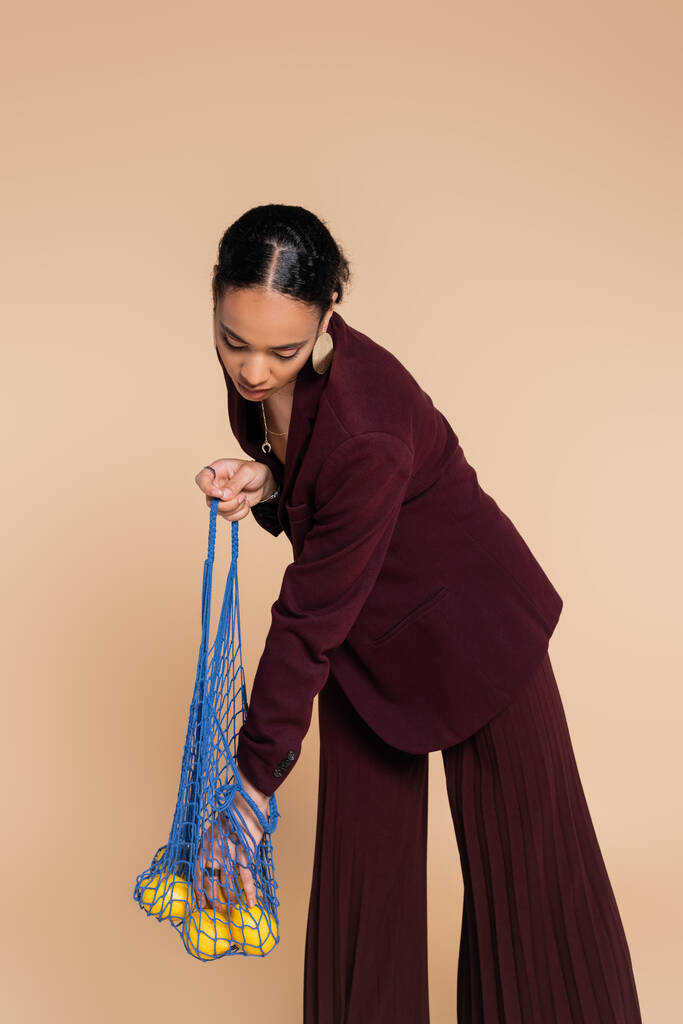 μοντέρνα αφροαμερικάνικη γυναίκα σε μπορντό κοστούμι κρατώντας τσάντα δίχτυ με λεμόνια που απομονώνονται σε μπεζ   - Φωτογραφία, εικόνα