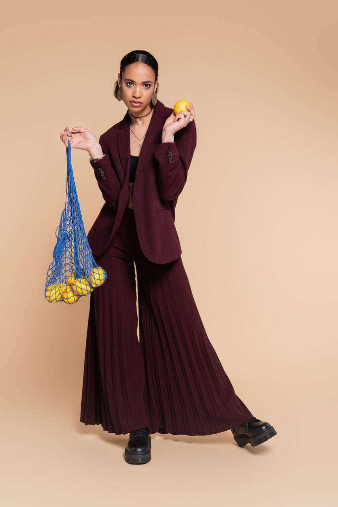 longueur totale du modèle afro-américain élégant en pantalon large bordeaux et veste tenant un sac en filet avec des citrons sur beige  - Photo, image
