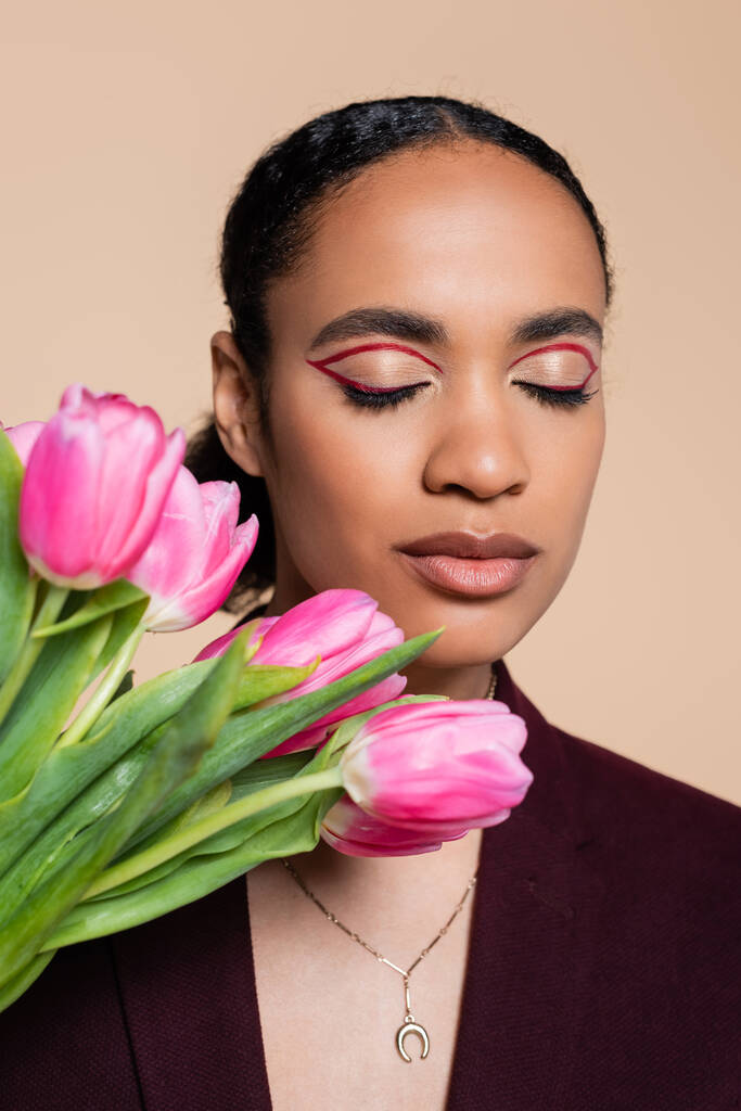encantadora mulher afro-americana com olhos fechados posando com tulipas rosa isoladas em bege  - Foto, Imagem