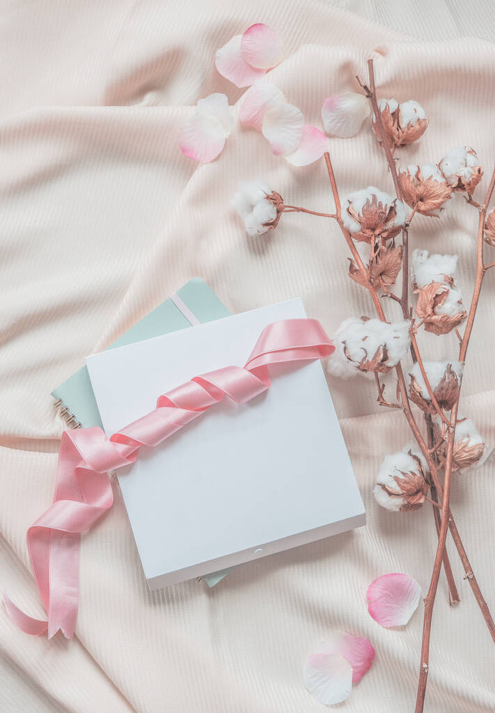 Stile di vita estetico con quaderno, scatola bianca e nastro rosa su sfondo tessuto beige con rami di cotone e petali rosa. Vista dall'alto. Bellezza - Foto, immagini