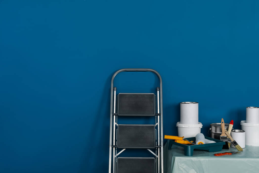 Escada dobrável metálica e ferramentas de pintura na mesa perto da parede azul, espaço para texto - Foto, Imagem