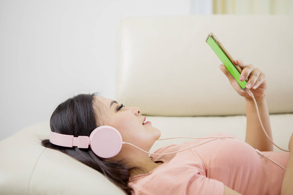 Klidná dívka v moderních bezdrátových sluchátek sedět relaxovat na pohodlném gauči poslouchat hudbu při pohledu na její smartphone - Fotografie, Obrázek