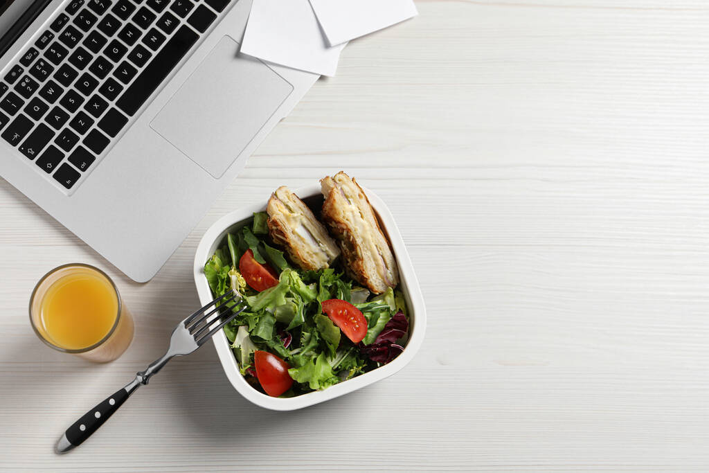 Container mit leckerem Essen, Laptop, Gabel und Saftglas auf weißem Holztisch, flache Liege mit Platz für Text. Geschäftsessen - Foto, Bild