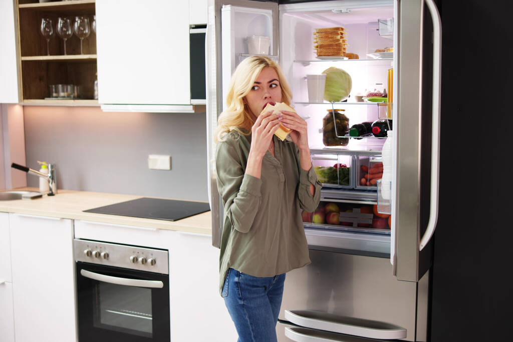Donna affamata che mangia sandwich al formaggio vicino al frigorifero aperto - Foto, immagini