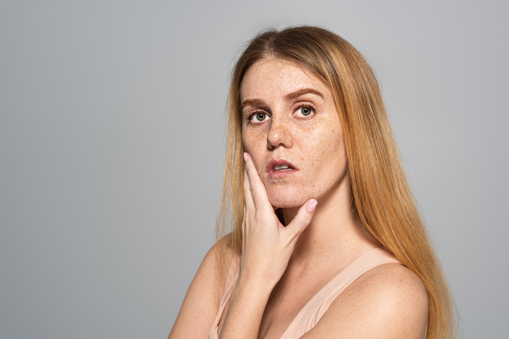 Junge Frau mit Sommersprossen im Gesicht berührt Haut isoliert auf grau  - Foto, Bild