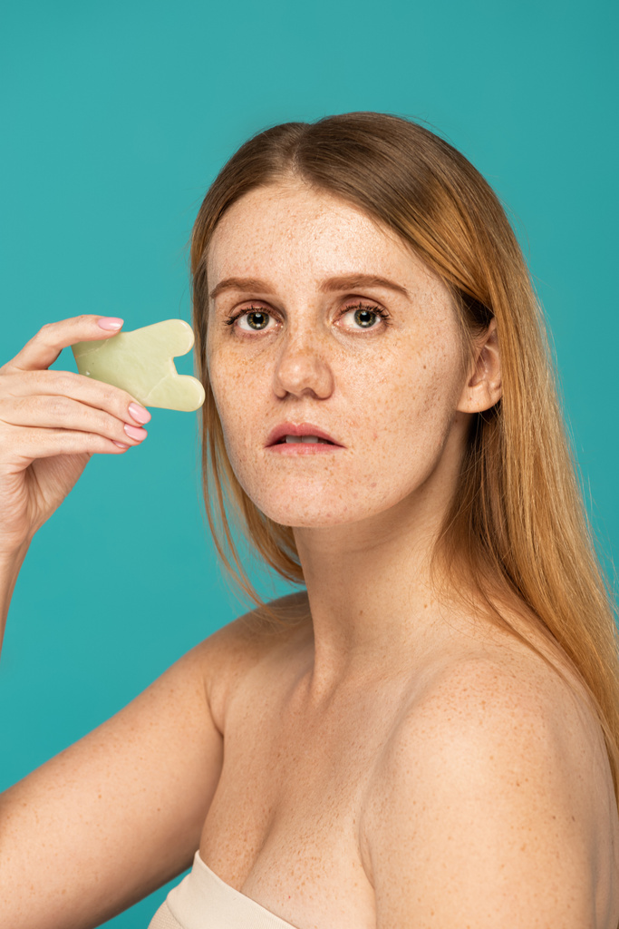 jonge vrouw met sproeten met behulp van gezicht scrapper geïsoleerd op turquoise - Foto, afbeelding