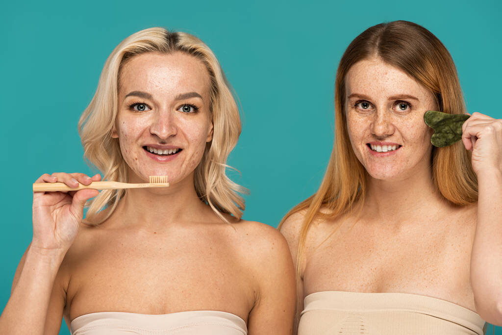 весела блондинка чистить зуби біля рудої моделі, використовуючи смітник для обличчя ізольовано на бірюзовому
  - Фото, зображення